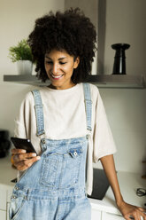 Smiling woman using cell phone in kitchen at home - VABF01835