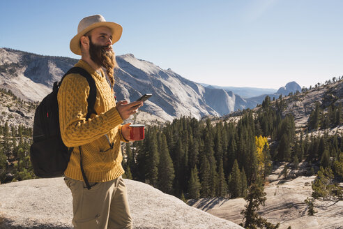 USA, Kalifornien, Yosemite-Nationalpark, Wanderer mit Smartphone und Becher in der Hand - KKAF03018