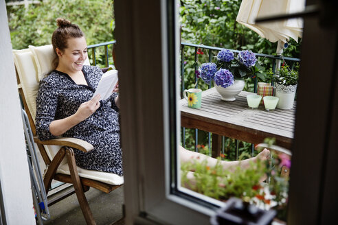 Lächelnde junge Frau sitzt auf dem Balkon und liest ein Buch - JATF01072