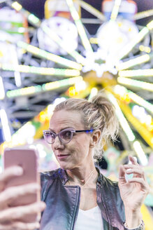 Porträt einer reifen Frau, die ein Selfie vor einem Riesenrad auf dem Jahrmarkt macht - FBAF00173