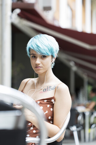 Porträt einer jungen Frau mit blau gefärbtem Haar, die in einem Straßencafé sitzt und mit Kopfhörern Musik hört, lizenzfreies Stockfoto