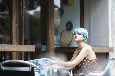 Porträt einer jungen Frau mit blau gefärbtem Haar, die in einem Straßencafé sitzt und mit Kopfhörern Musik hört - ERRF00153