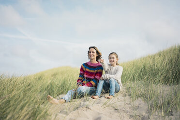 Mother and daughter sitting on a beach dune, girl pointing at distance, smiling - MOEF01580