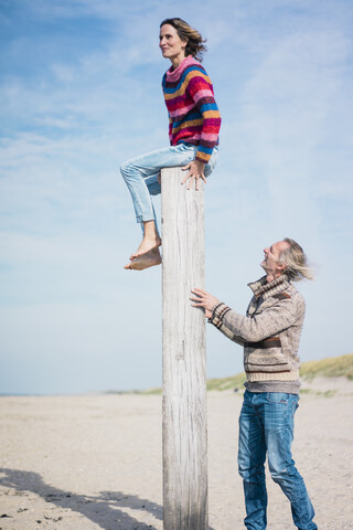Älterer Mann hilft Frau beim Klettern auf einen Holzpfahl am Strand, lizenzfreies Stockfoto