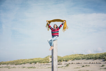 Ältere Frau sitzt auf einem Holzpfahl am Strand und hält ein Tuch in den Wind - MOEF01618