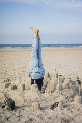 Ältere Frau macht einen Kopfstand am Strand in einer Sandburg, lizenzfreies Stockfoto