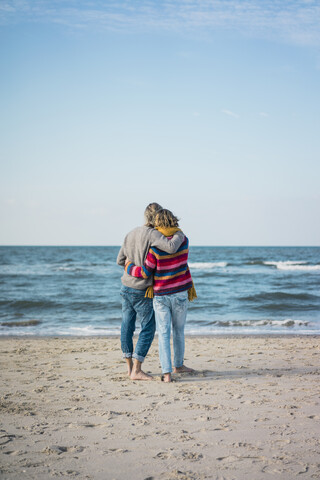 ein hübsches Paar, das am Strand steht, die Arme um sich legt und auf das Meer schaut, lizenzfreies Stockfoto