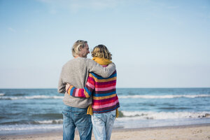 ein hübsches Paar, das am Strand steht, die Arme um sich legt und auf das Meer schaut - MOEF01635