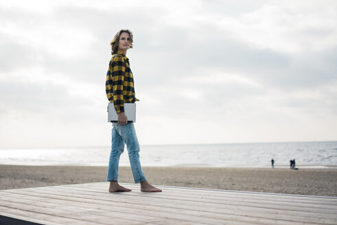 Ältere Frau steht auf der Promenade am Strand und hält einen Laptop - MOEF01643