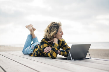 Mature woman lying on boardwalk at the beach, using laptop - MOEF01645