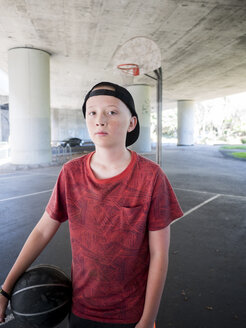 Porträt eines selbstbewussten Jungen, der einen Ball auf einem Basketballplatz hält - CAVF56030