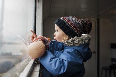 Seitenansicht eines überraschten Mädchens mit Spielzeug, das durch ein Fenster schaut - CAVF56131