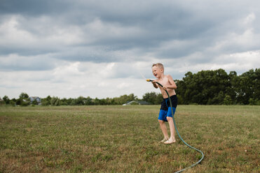 In voller Länge von shirtless spielerischen Jungen spielen mit Gartenschlauch auf grasbewachsenen Feld gegen bewölkten Himmel im Hinterhof - CAVF56152