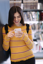 Lächelnde Frau, die ein Mobiltelefon benutzt, während sie in einer Bibliothek steht - CAVF56167