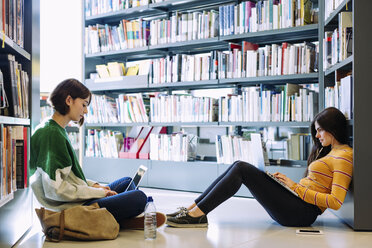 Freundinnen, die in einer Bibliothek auf dem Boden sitzend mit einem Laptop lernen - CAVF56182