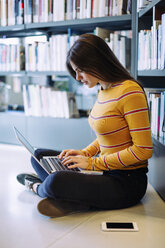 Seitenansicht einer Frau, die einen Laptop benutzt, während sie in einer Bibliothek auf dem Boden sitzt - CAVF56184