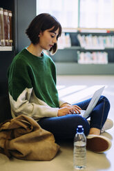 Frau, die einen Laptop benutzt, während sie in einer Bibliothek auf dem Boden sitzt - CAVF56185