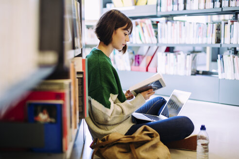 Seitenansicht einer Frau mit Laptop, die ein Buch liest, während sie in einer Bibliothek auf dem Boden sitzt - CAVF56188