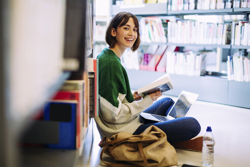 Porträt einer Frau mit Laptop, die in einer Bibliothek auf dem Boden sitzend ein Buch liest - CAVF56189