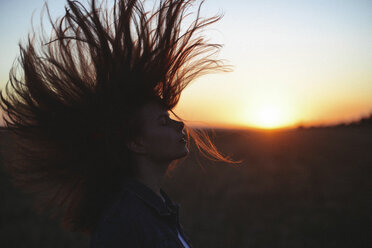 Side view of woman with eyes closed tossing hair while standing against sky during sunset - CAVF56213