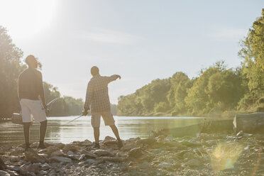 Rear view of male friends with fishing rods standing at lakeshore against sky during sunny day - CAVF56283