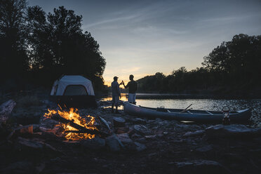 Rückansicht von männlichen Freunden, die auf dem Campingplatz am See bei Sonnenuntergang mit Getränken anstoßen - CAVF56290