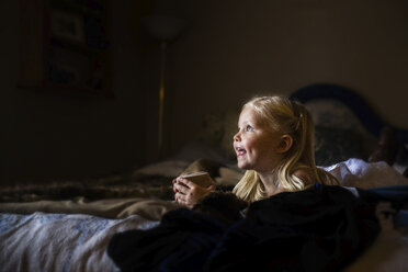 Mädchen hält Kekse in der Hand, während sie zu Hause auf dem Bett liegt - CAVF56300