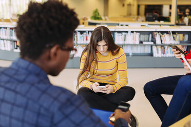 Friends using smart phones while sitting in library - CAVF56318