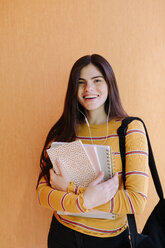 Portrait of smiling woman with books standing against orange wall in library - CAVF56327