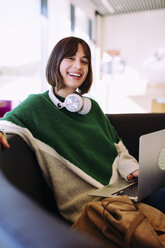 Porträt einer lächelnden Frau, die einen Laptop benutzt und auf einem Sofa in einer Bibliothek sitzt - CAVF56343