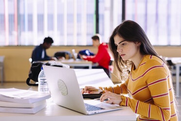 Seitenansicht einer Frau, die einen Laptop benutzt, während männliche Freunde im Hintergrund in einer Bibliothek lernen - CAVF56346