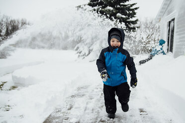 Glücklicher Junge in warmer Kleidung, der auf einem schneebedeckten Feld im Hof läuft - CAVF56392