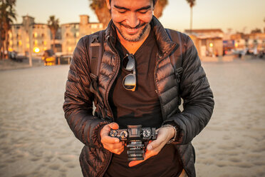 Smiling man wearing jacket while holding camera at beach - CAVF56395
