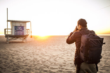 Rückansicht eines Rucksacktouristen, der eine Rettungsschwimmerhütte mit einer Kamera am Strand bei Sonnenuntergang fotografiert - CAVF56396