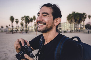 Close-up of smiling backpacker holding camera while looking away at beach - CAVF56398