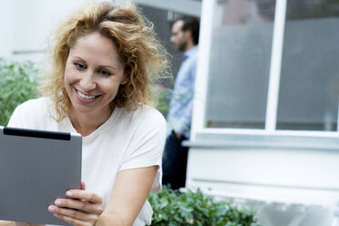 Lachende Frau, die im Garten sitzend an ihrem digitalen Tablet arbeitet - HHLMF00560