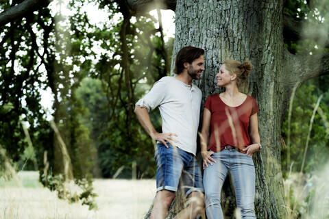 Glückliches Paar lehnt an einem Baum und lächelt sich an, lizenzfreies Stockfoto