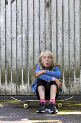 Portrait of girl sitting on skateboard in front of wooden wall sticking out tongue - JFEF00918