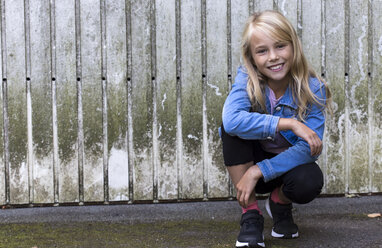 Portrait of smiling blond girl crouching in front of wooden wall - JFEF00925