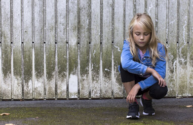 Porträt eines ernsten blonden Mädchens, das vor einer Holzwand hockt - JFEF00926