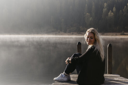 Schweiz, Engadin, Stazer See, lächelnde junge Frau sitzt auf einem Steg am Seeufer in der Morgensonne - LHPF00145