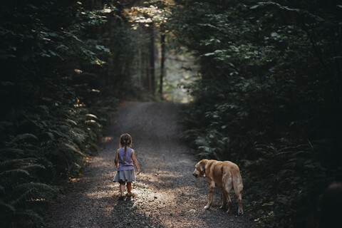 Rückansicht eines Mädchens mit Hund, das auf einem Feldweg inmitten von Pflanzen im Wald steht, lizenzfreies Stockfoto