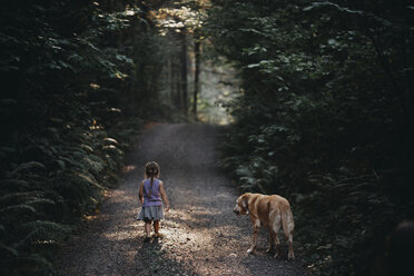 Rear view of girl with dog standing on dirt road amidst plants at forest - CAVF56452