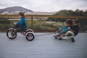 Boy pulling toy car with friends while riding tricycle on floorboard - CAVF56527