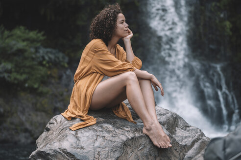 Nachdenkliche Frau schaut weg, während sie auf einem Felsen vor einem Wasserfall im Wald sitzt - CAVF56532