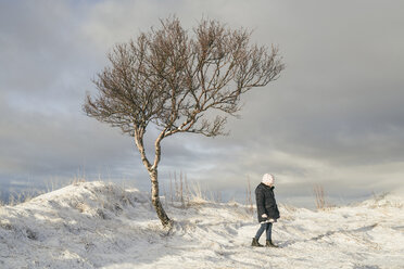 Mädchen in voller Länge an einem kahlen Baum auf einem verschneiten Feld stehend - CAVF56541