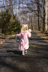 Rückansicht eines Mädchens mit Ostereiern und Stofftier, das auf einer Straße inmitten von Bäumen läuft - CAVF56550