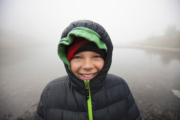 Porträt eines lächelnden Jungen in warmer Kleidung am See bei nebligem Wetter - CAVF56606