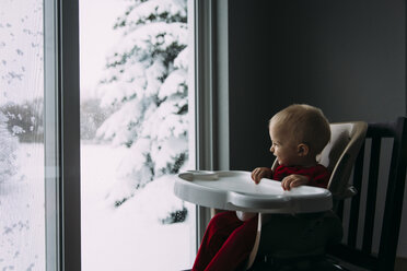 Glücklicher kleiner Junge, der durch das Fenster schaut, während er auf dem Hochstuhl zu Hause im Winter sitzt - CAVF56613
