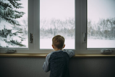 Rear view of boy looking through window while standing at home during winter - CAVF56614